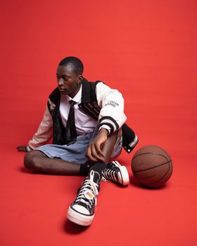 Young adult in trendy outfit sitting with basketball on a red backdrop.