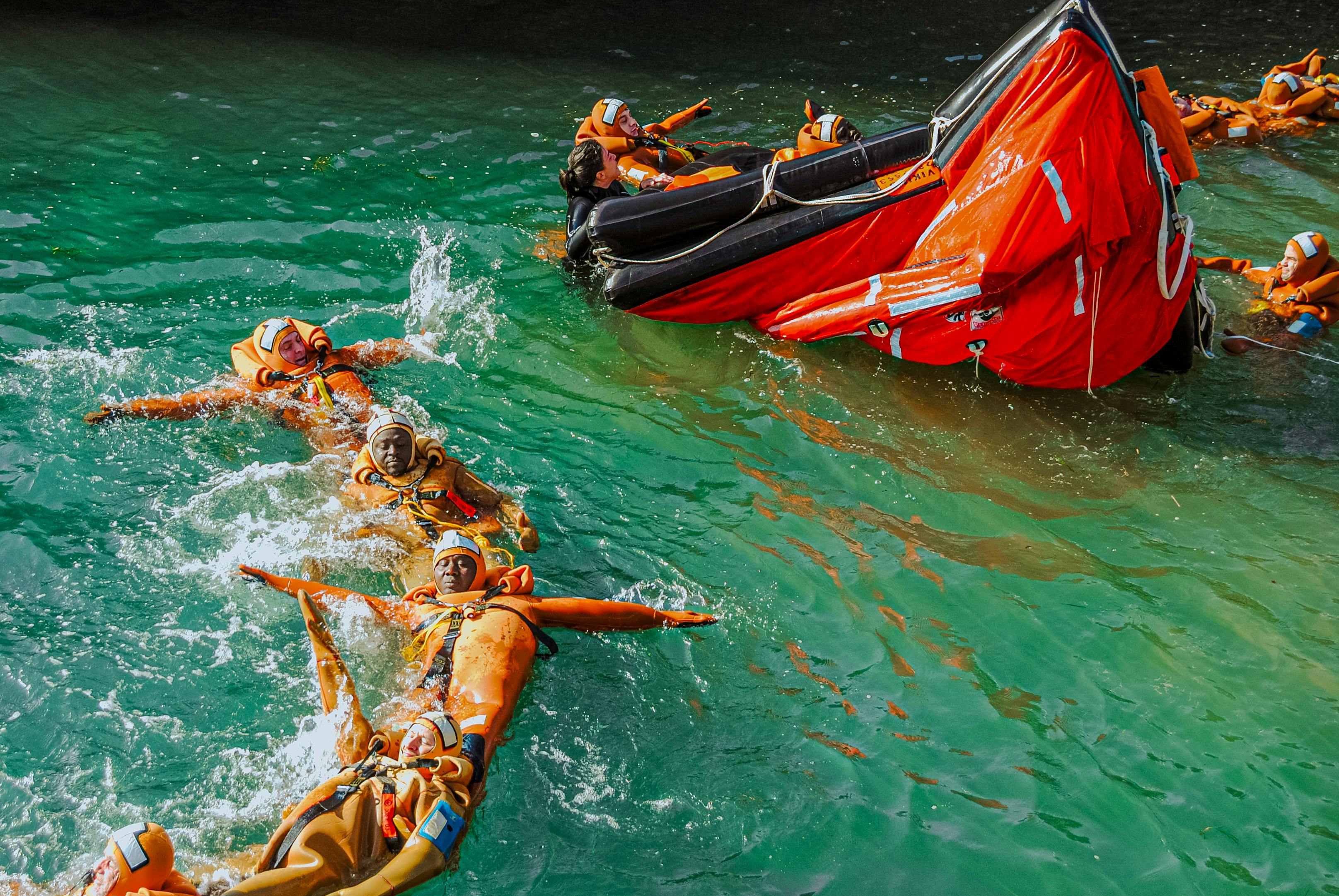 Group of adults practicing lifeboat safety drill in the water wearing life jackets.