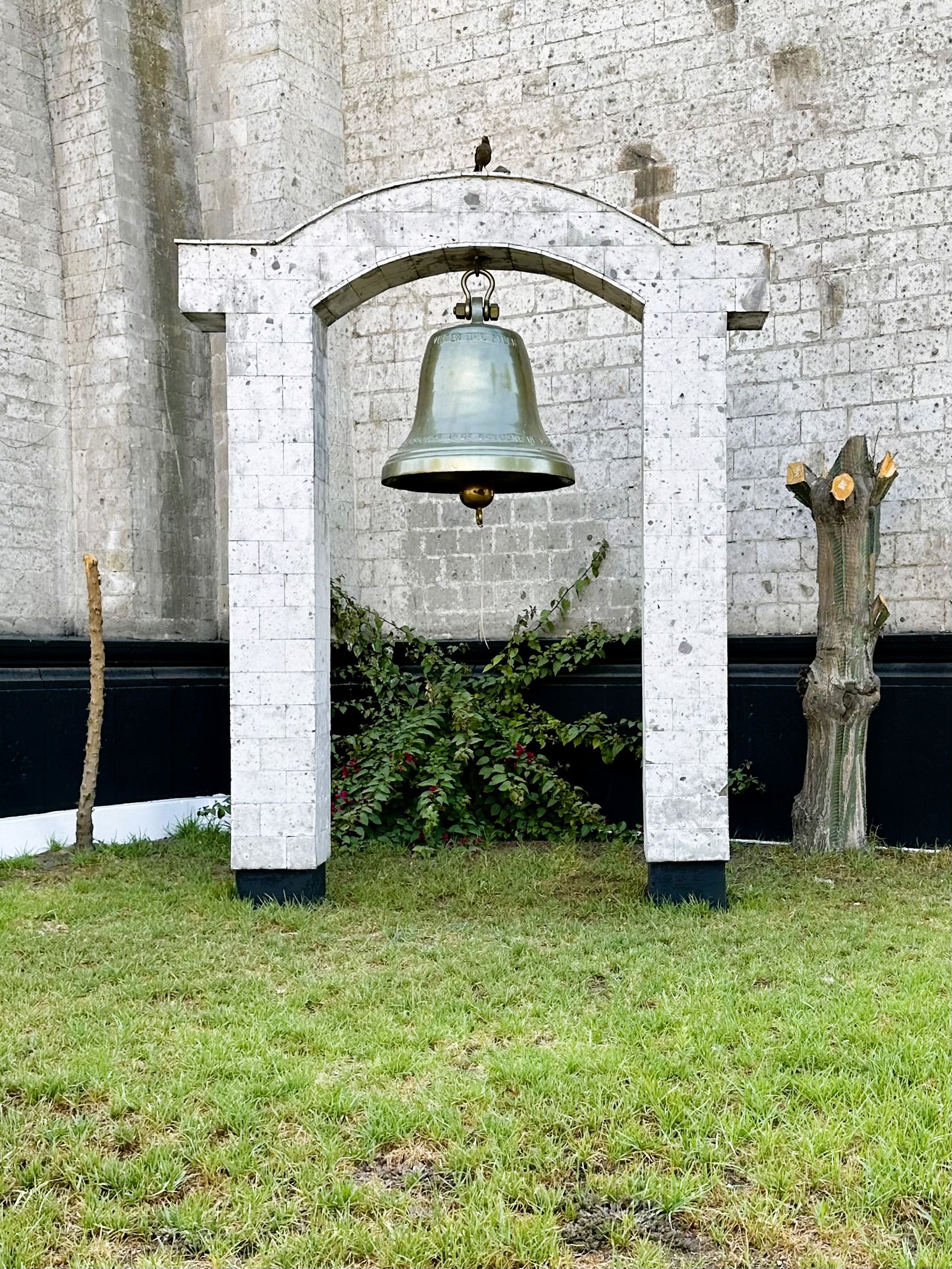 A large bronze bell hanging under a stone arch in a green courtyard against a brick wall.
