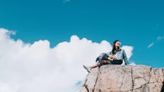 一位年轻女子幸福地坐在阳光明媚的蓝天下岩石上