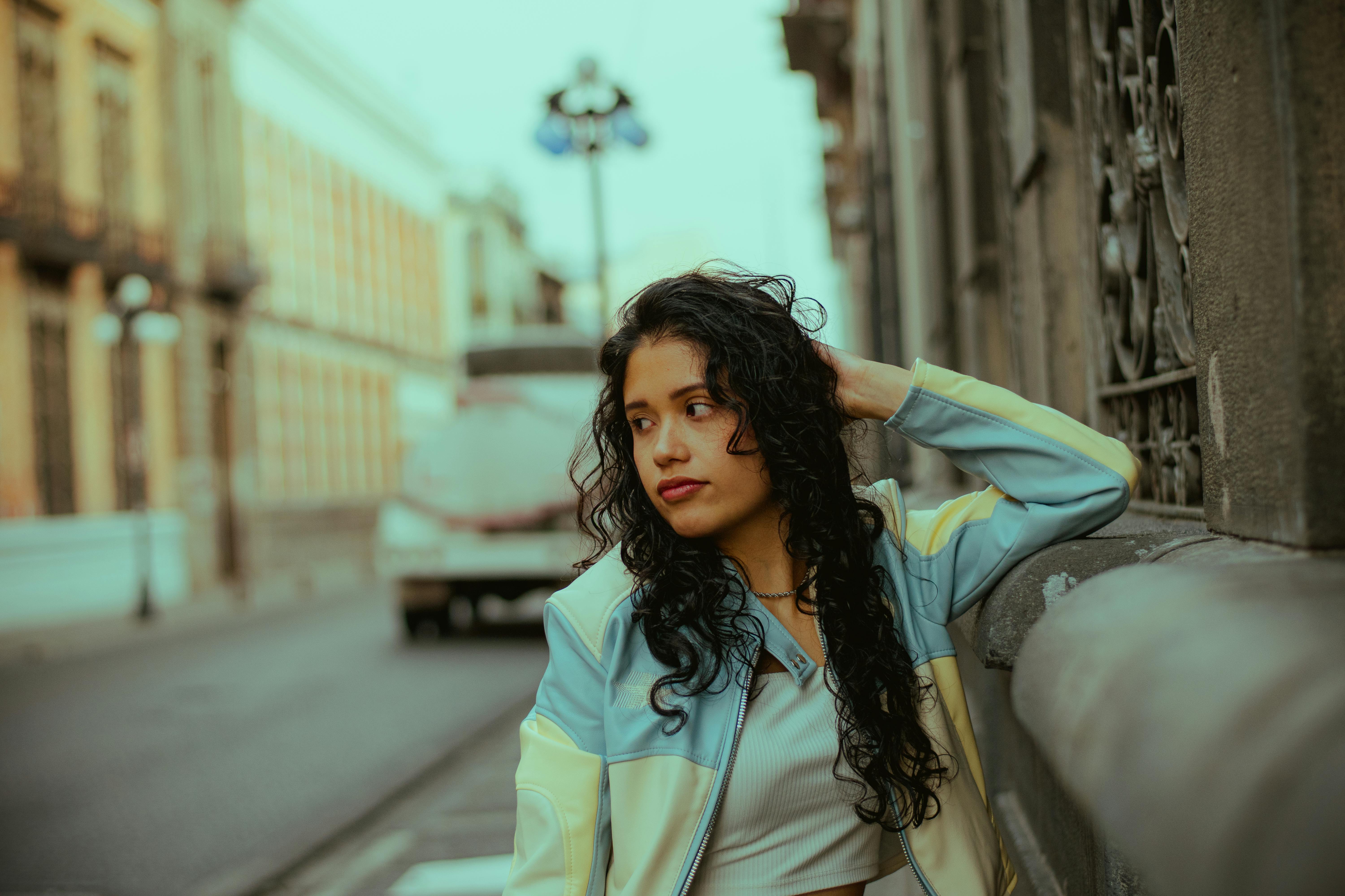 A young woman with curly hair poses outdoors, leaning against a stone wall in a city setting.
