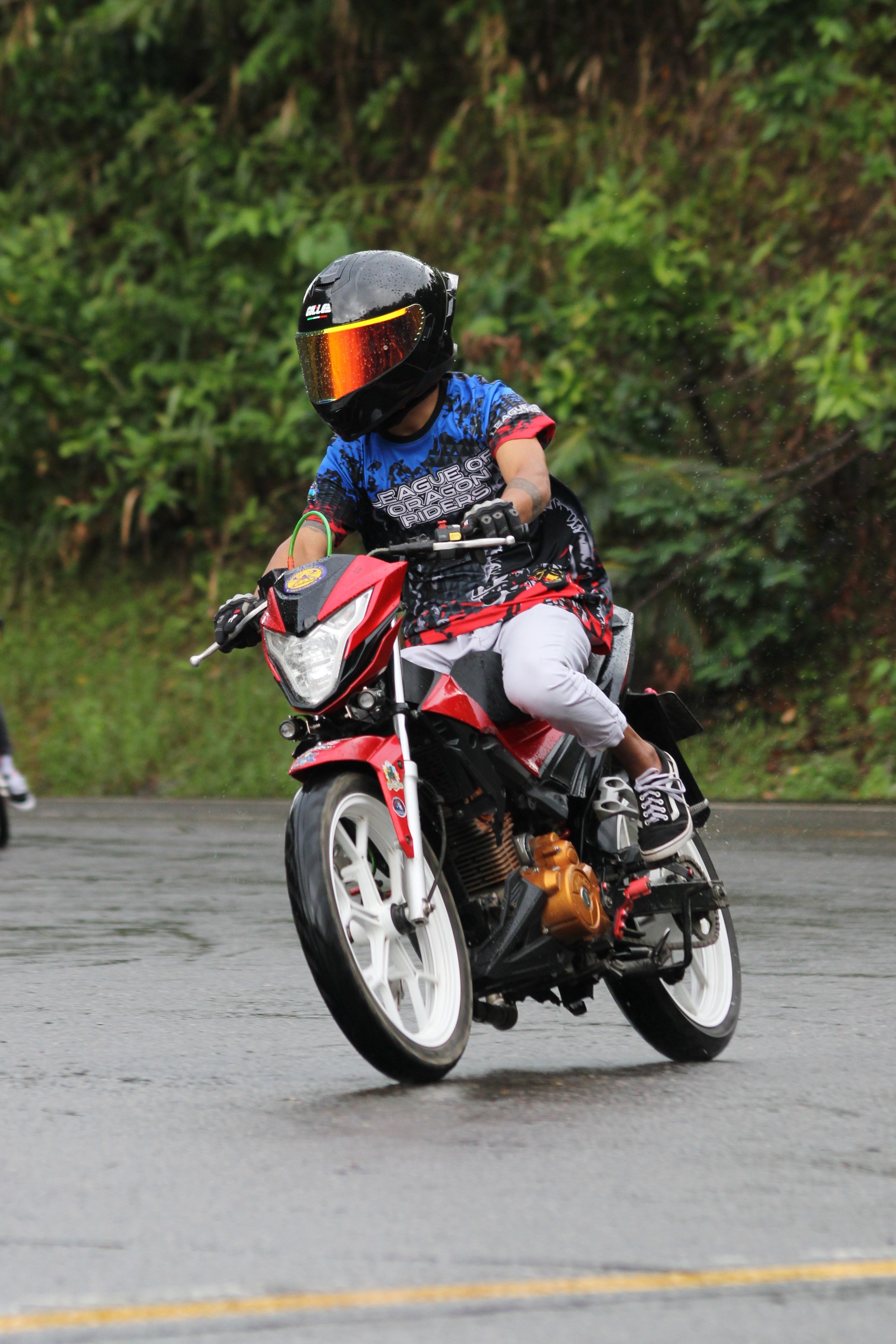 A motorcyclist performs a thrilling stunt on a wet road against a lush green backdrop.