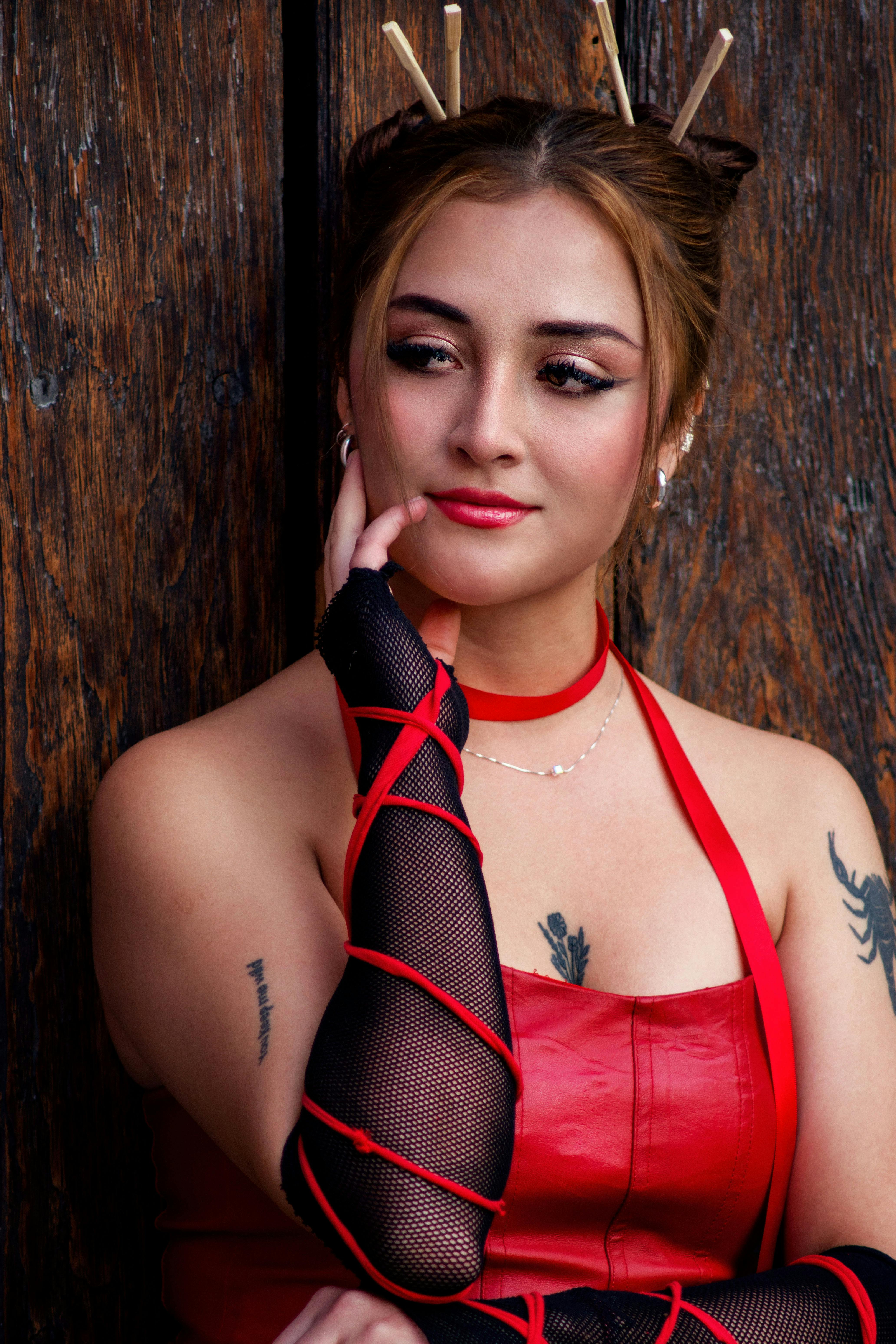A woman dressed in red with tattoos, standing against a wooden background.