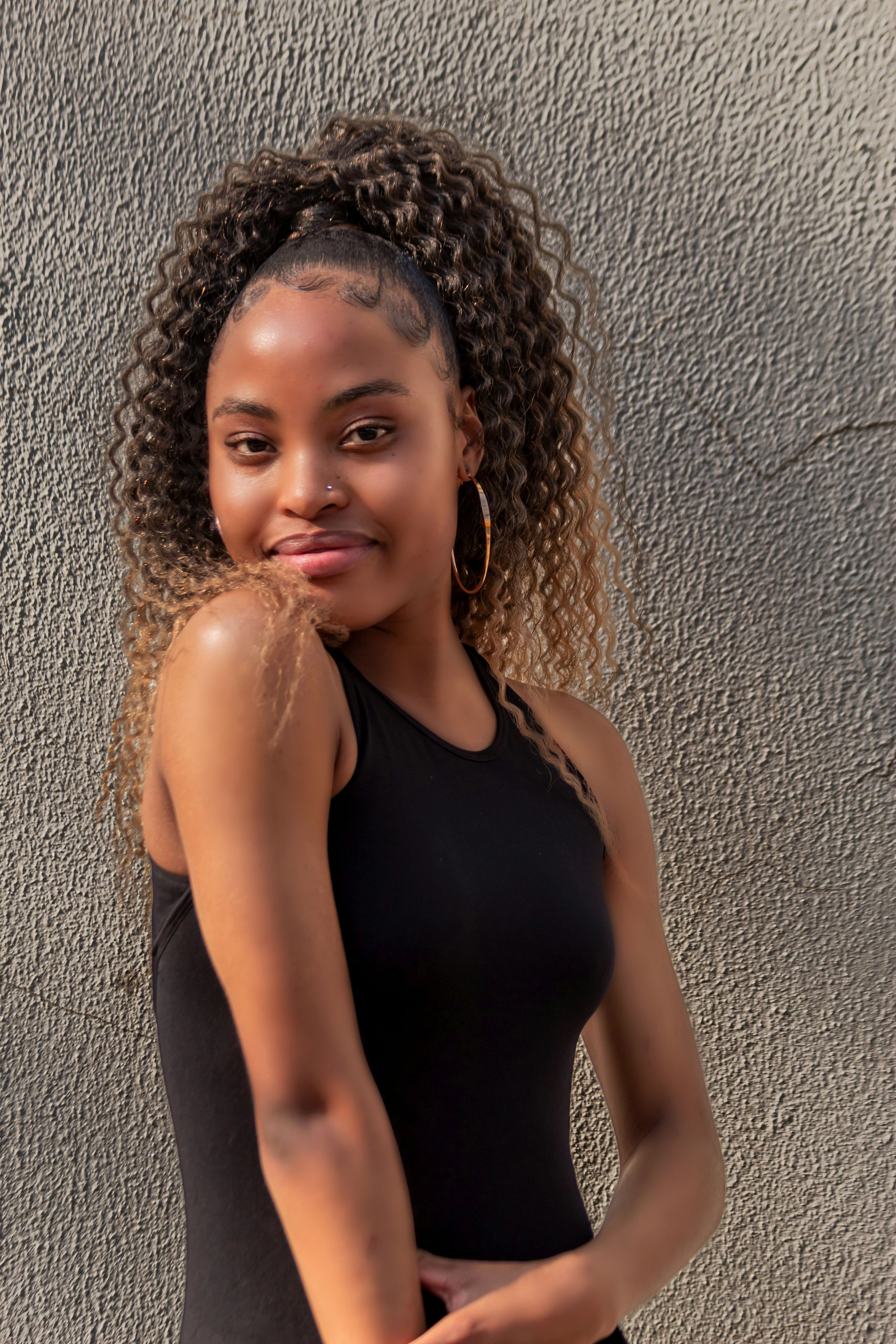 Portrait of a young woman in a black tank top smiling against a textured wall.