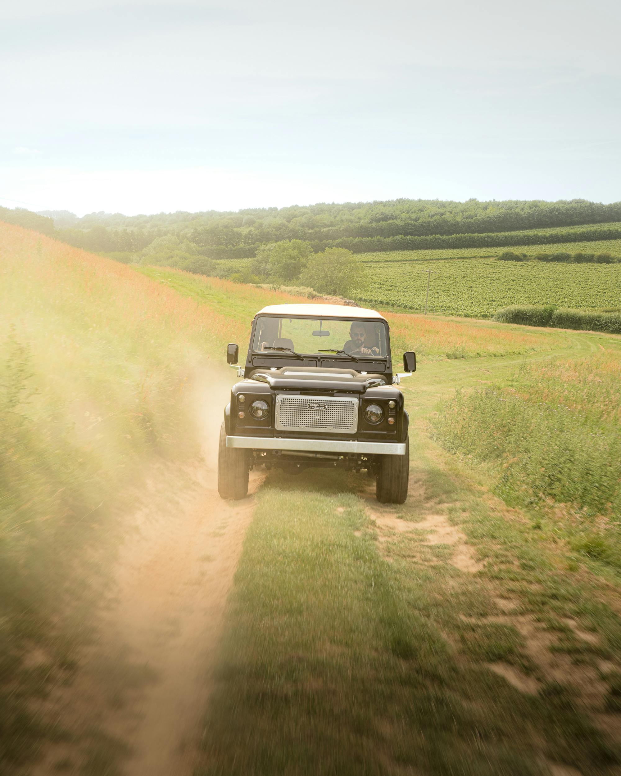 A rugged vehicle driving through a dusty country road amidst lush green fields under a clear sky.