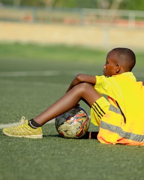 A young soccer player in a yellow jersey takes a break on the grass field with a soccer ball.