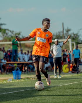A young soccer player in orange jersey controls the ball mid-game outdoors.