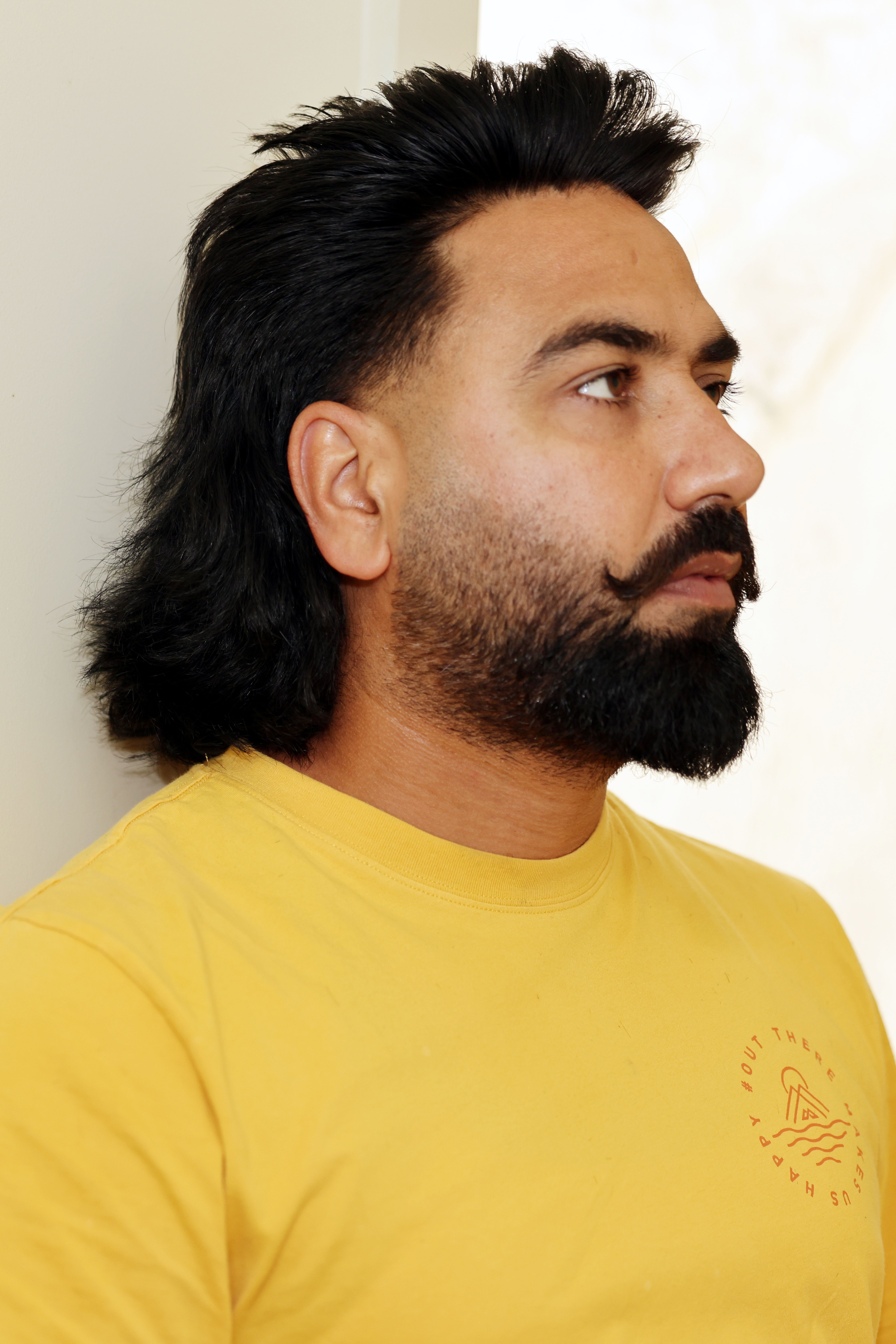 Side profile portrait of a man with a stylish beard and long hair wearing a yellow shirt.