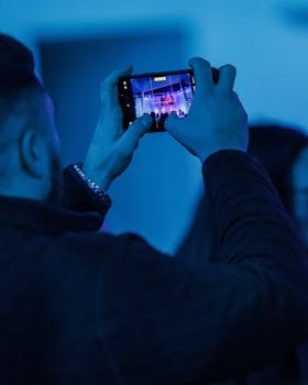 Man records a live music performance on smartphone under blue lighting.