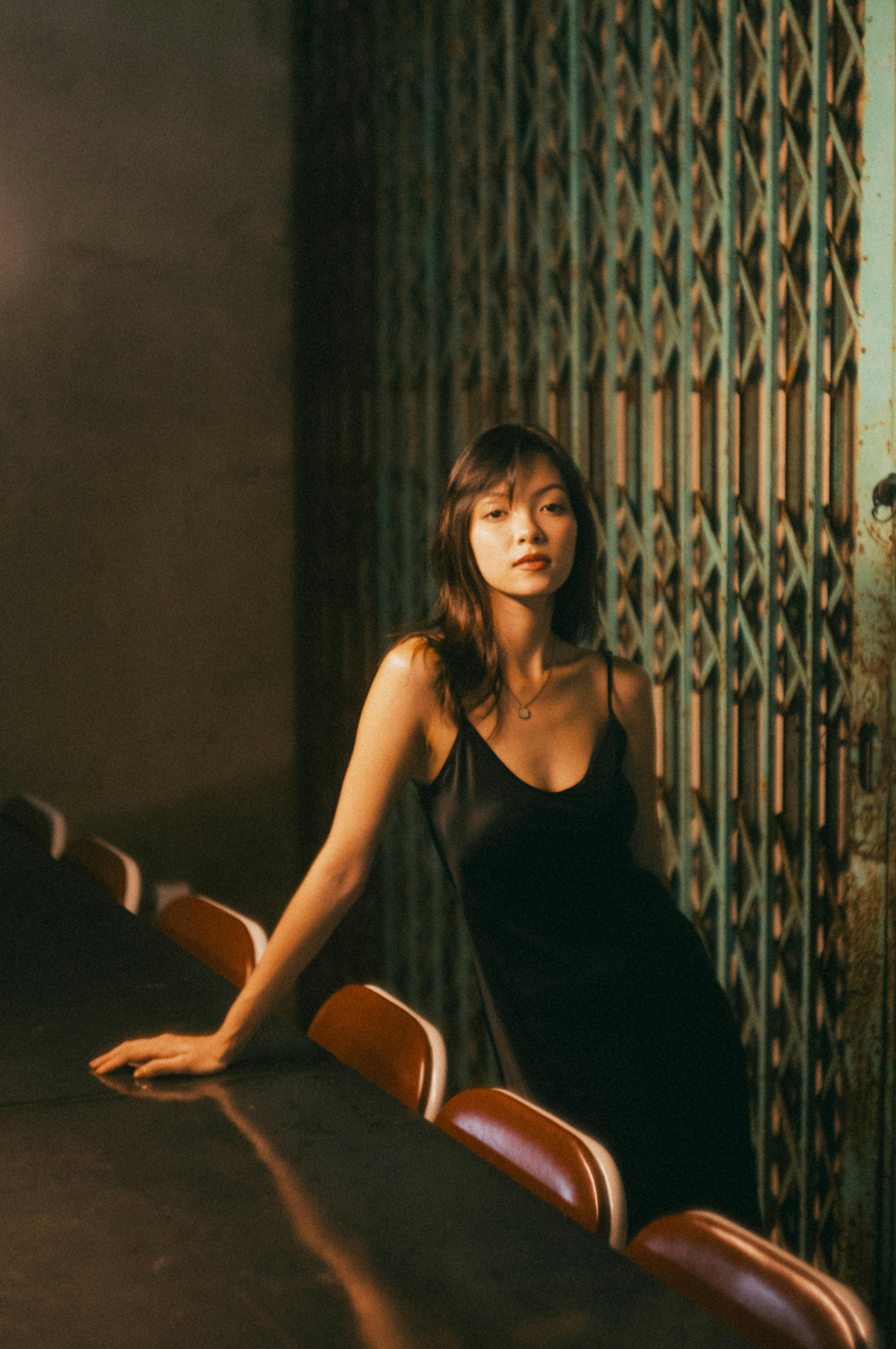 Portrait of a woman in a black dress leaning on a table in a moody, dimly lit environment.