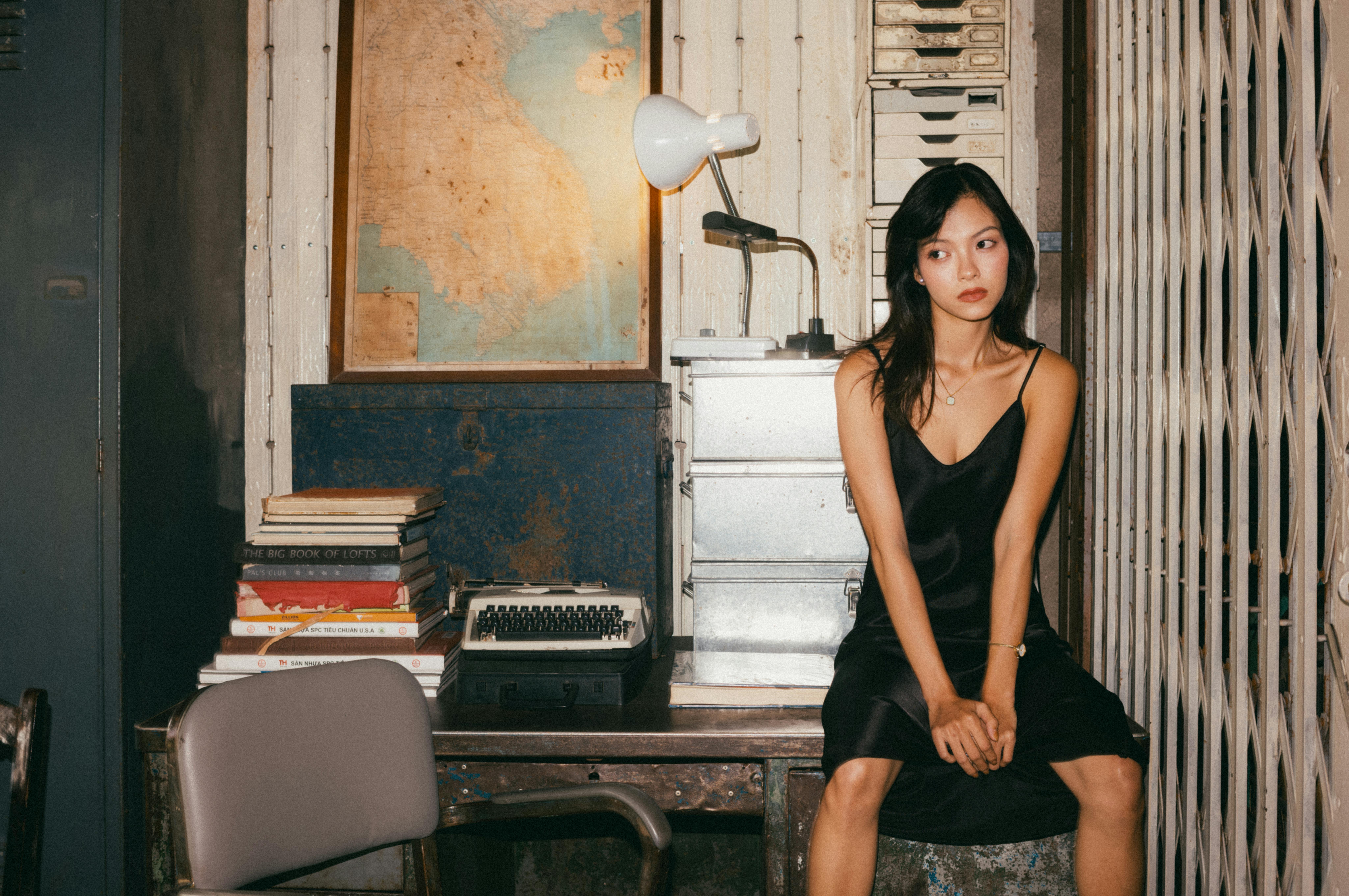 Young woman in black dress sitting pensively in a vintage office setting with a typewriter.