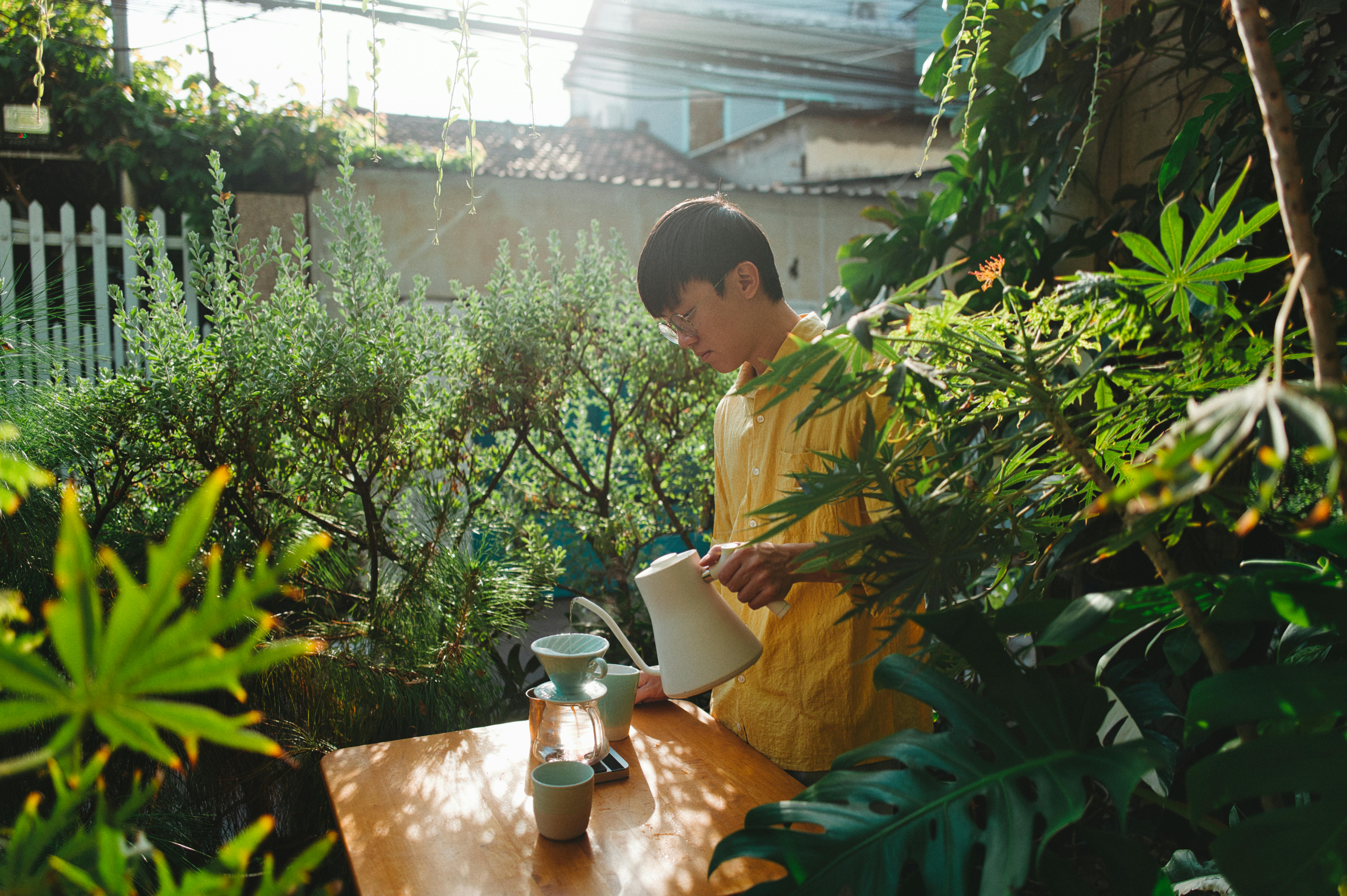 A man prepares coffee in a vibrant outdoor garden setting, surrounded by lush greenery.