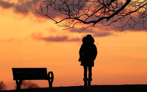Free A serene silhouette of a person standing next to a bench against a vibrant sunrise sky. Stock Photo