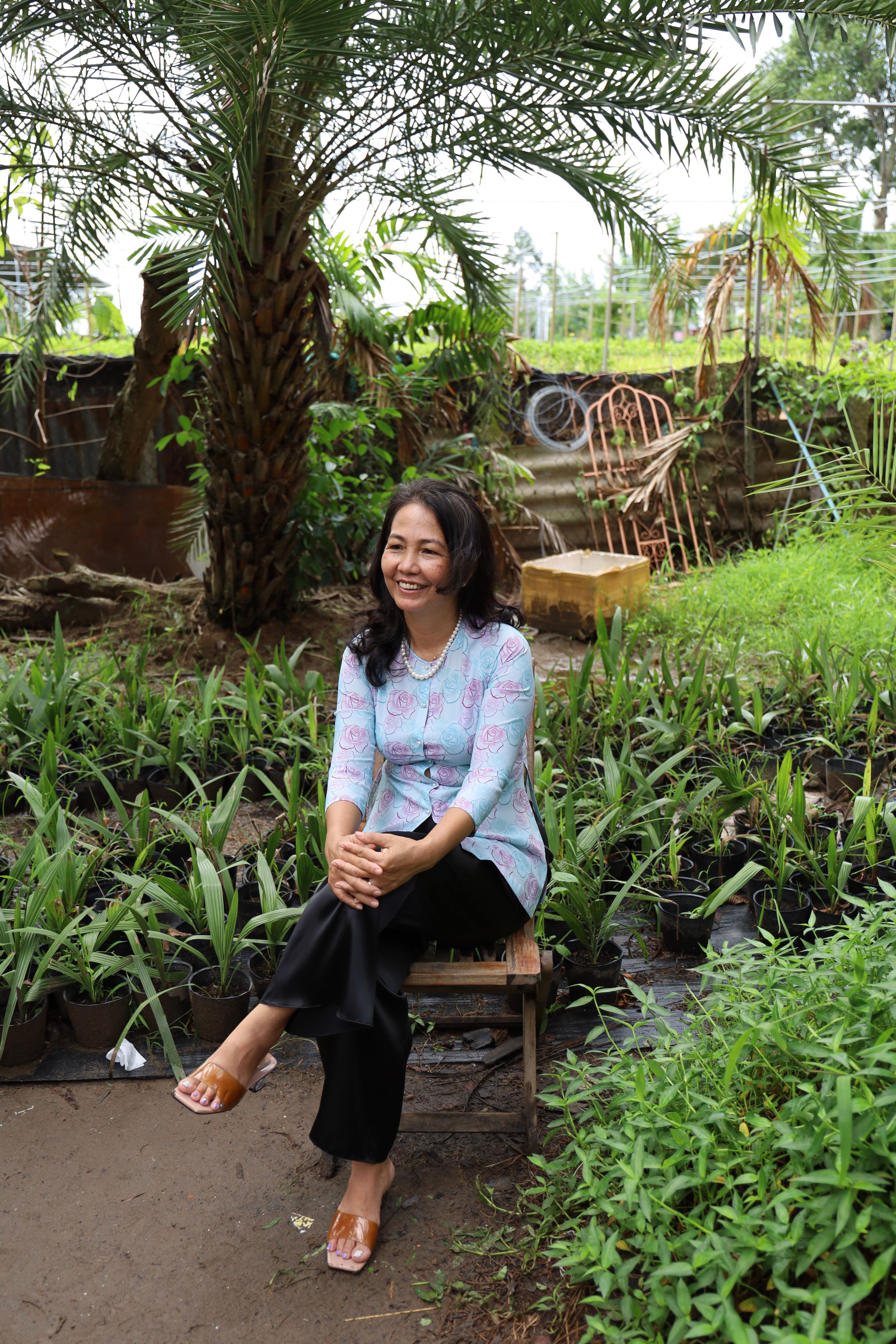 Woman sitting amidst lush plants in a tropical garden, smiling warmly.