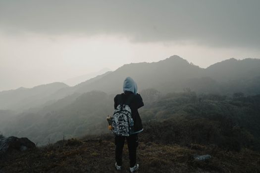 A lone hiker in a hoodie stands on a mountain, gazing at mist-covered peaks in a moody landscape.