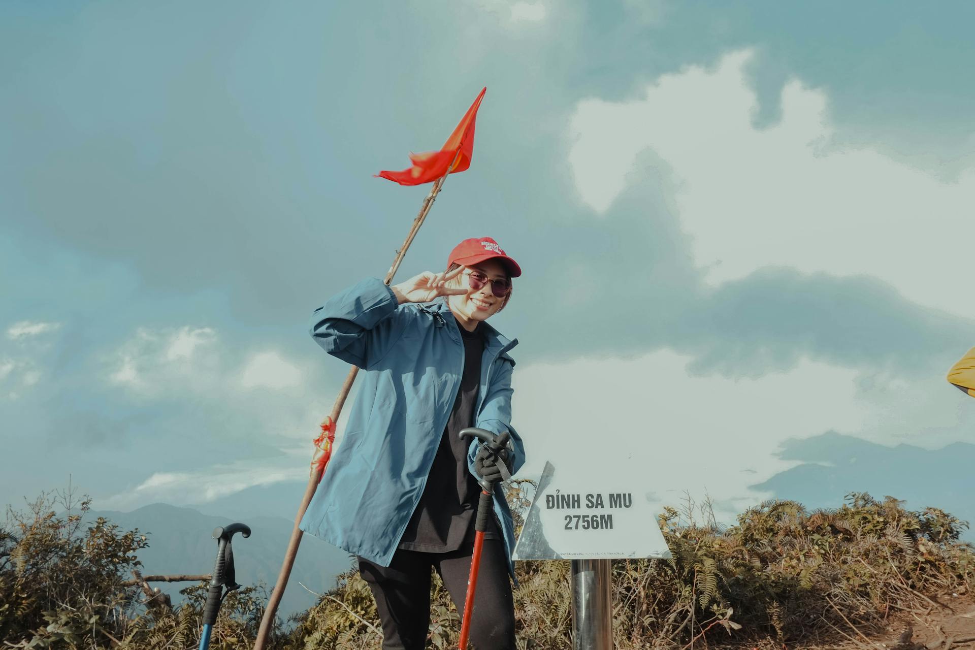 A person poses triumphantly at Đỉnh Sa Mu peak holding a red flag. Scenic mountain view.