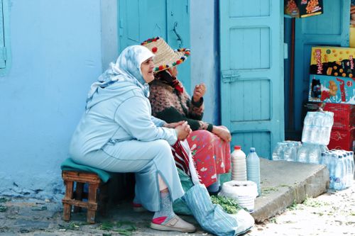 Free Two women in traditional attire chatting outside a blue Moroccan market. Stock Photo