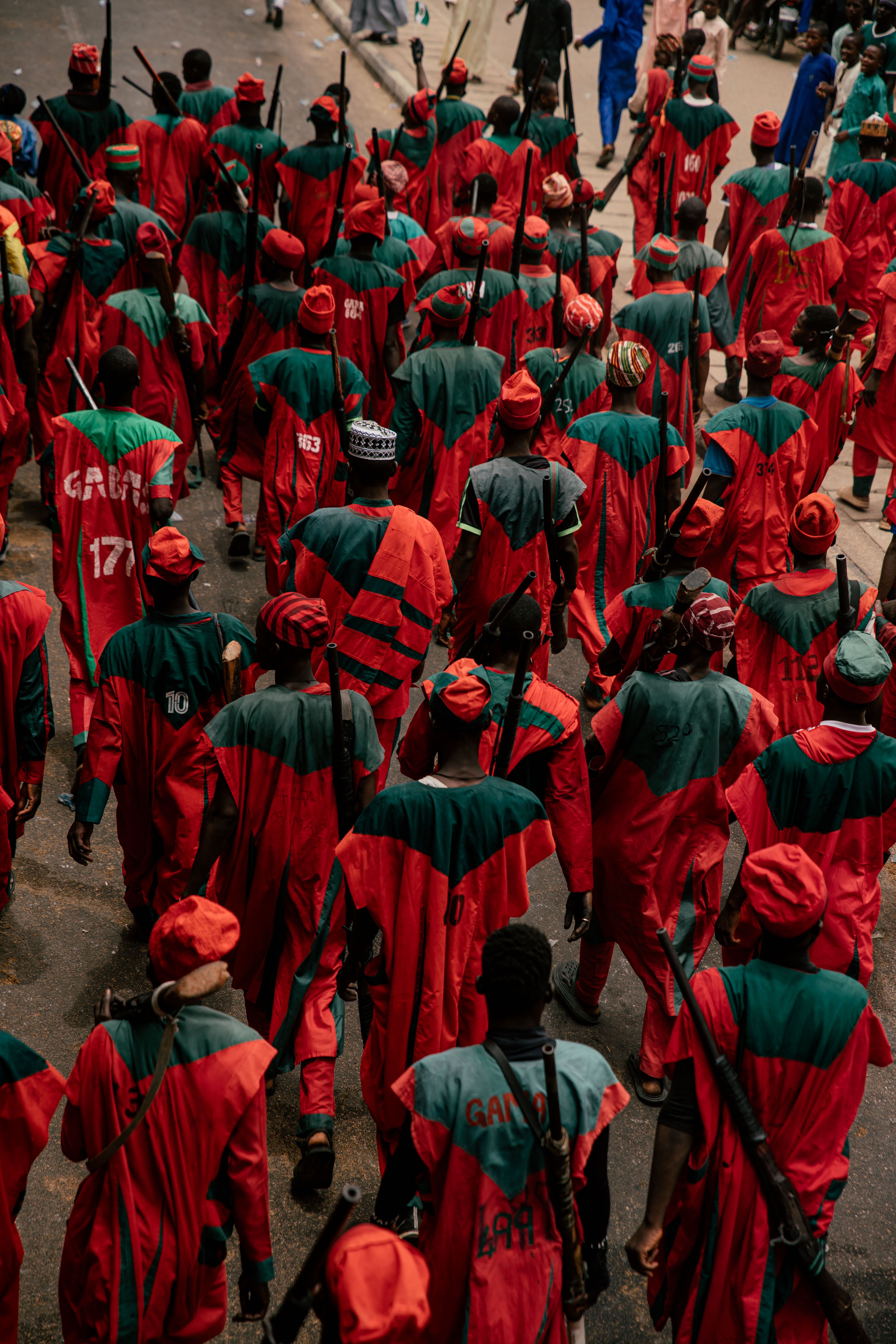 Street procession with people in colorful traditional attire holding walking sticks.