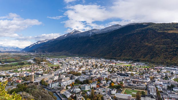 A stunning aerial view of a Swiss town surrounded by mountains under a cloudy sky, showcasing urban and natural beauty.