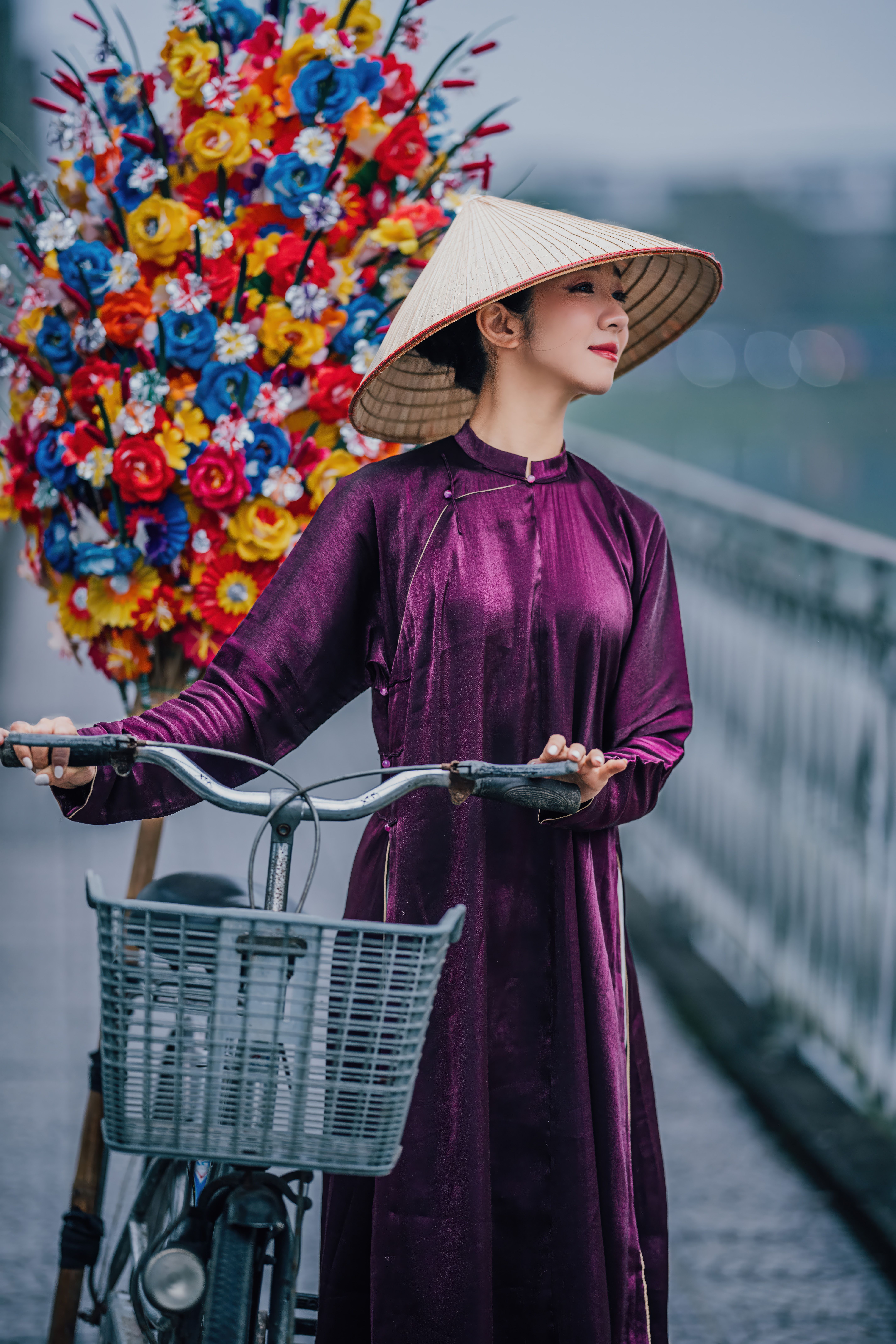 Woman in traditional Ao Dai with bicycle on bridge beside vibrant flower display.