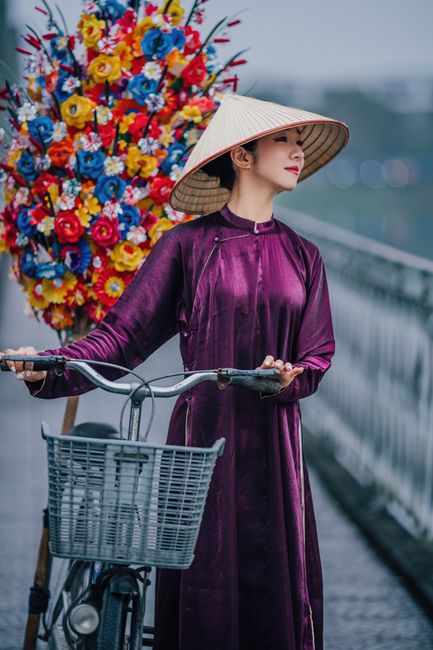 Woman in traditional Ao Dai with bicycle on bridge beside vibrant flower display.
