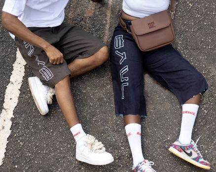 Two individuals in trendy streetwear rest on an urban road, showcasing embroidered shorts and colorful sneakers.