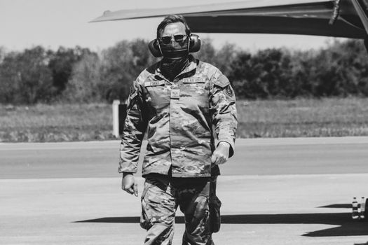 A military officer in camouflage gear and earmuffs stands on an airfield in black and white.