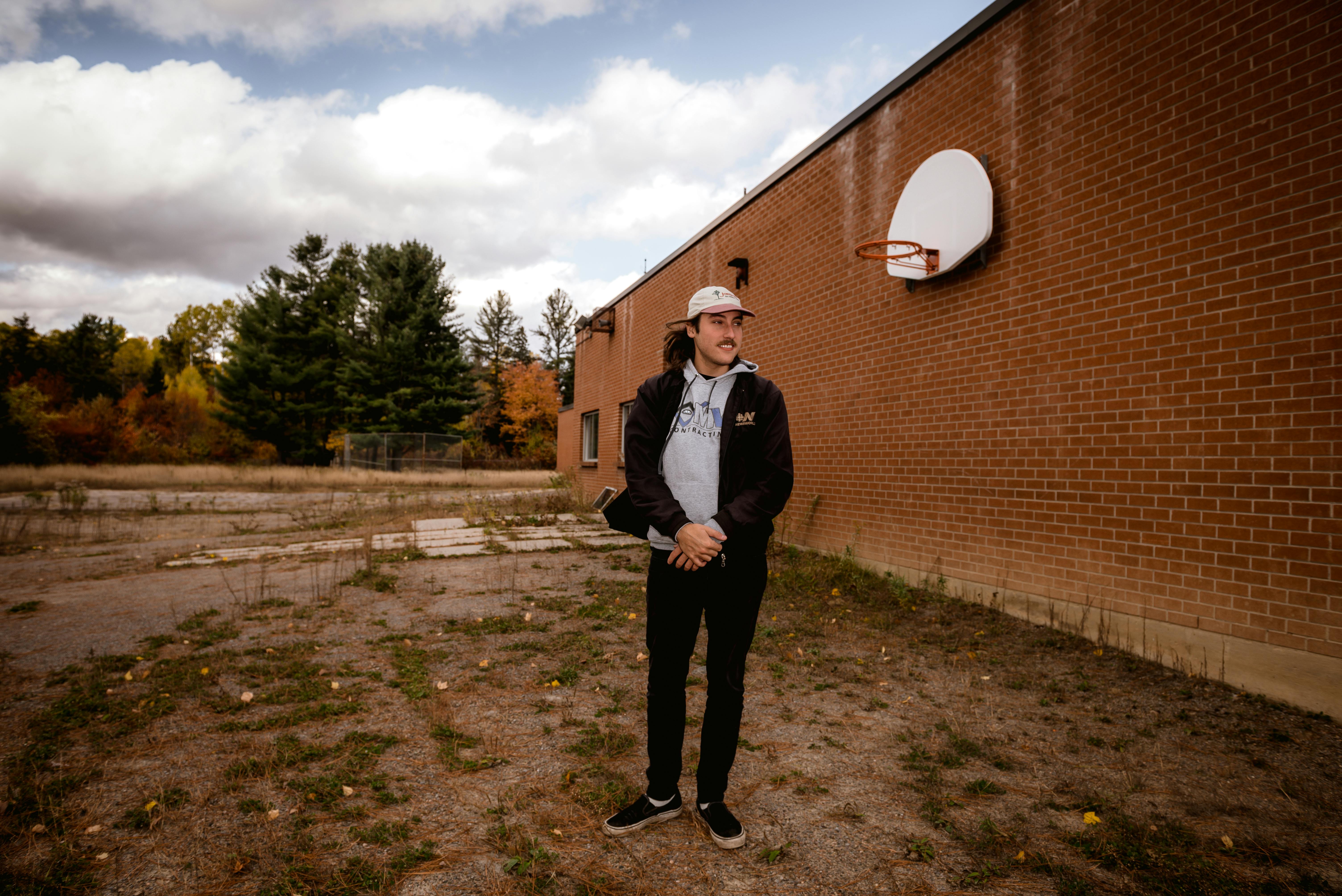 一名穿着休闲装的年轻人站在砖墙边，手里拿着篮球筐