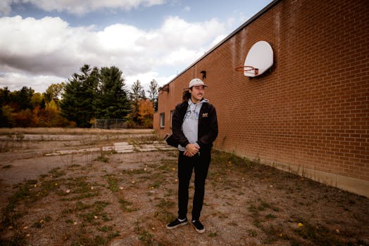 一名穿着休闲装的年轻人站在砖墙边，手里拿着篮球筐