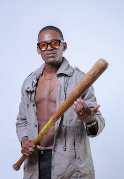 Confident man wearing glasses and jacket holding a baseball bat. Studio shot with minimal background.