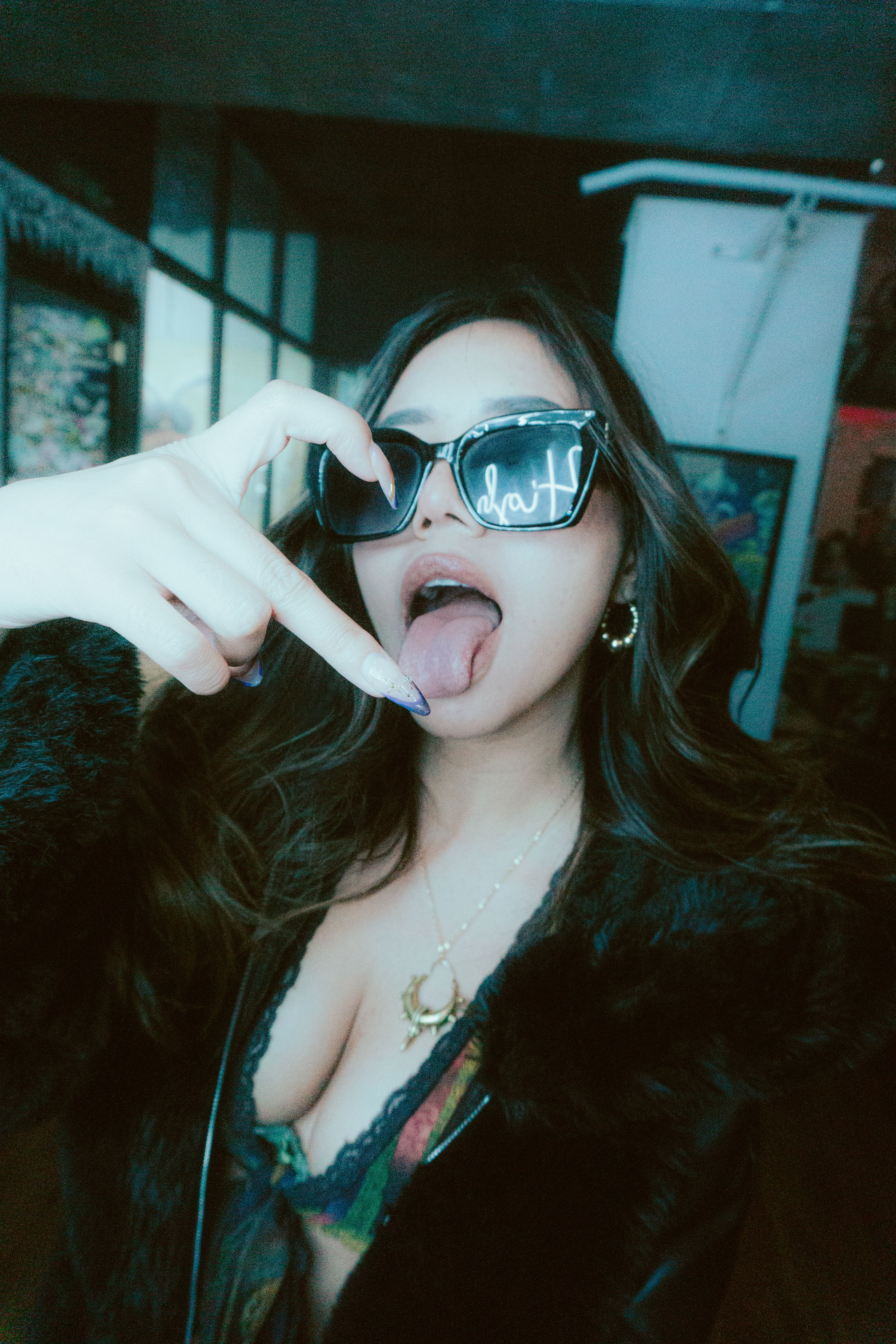 Stylish woman in sunglasses and fur coat posing playfully indoors with a trendy vibe.