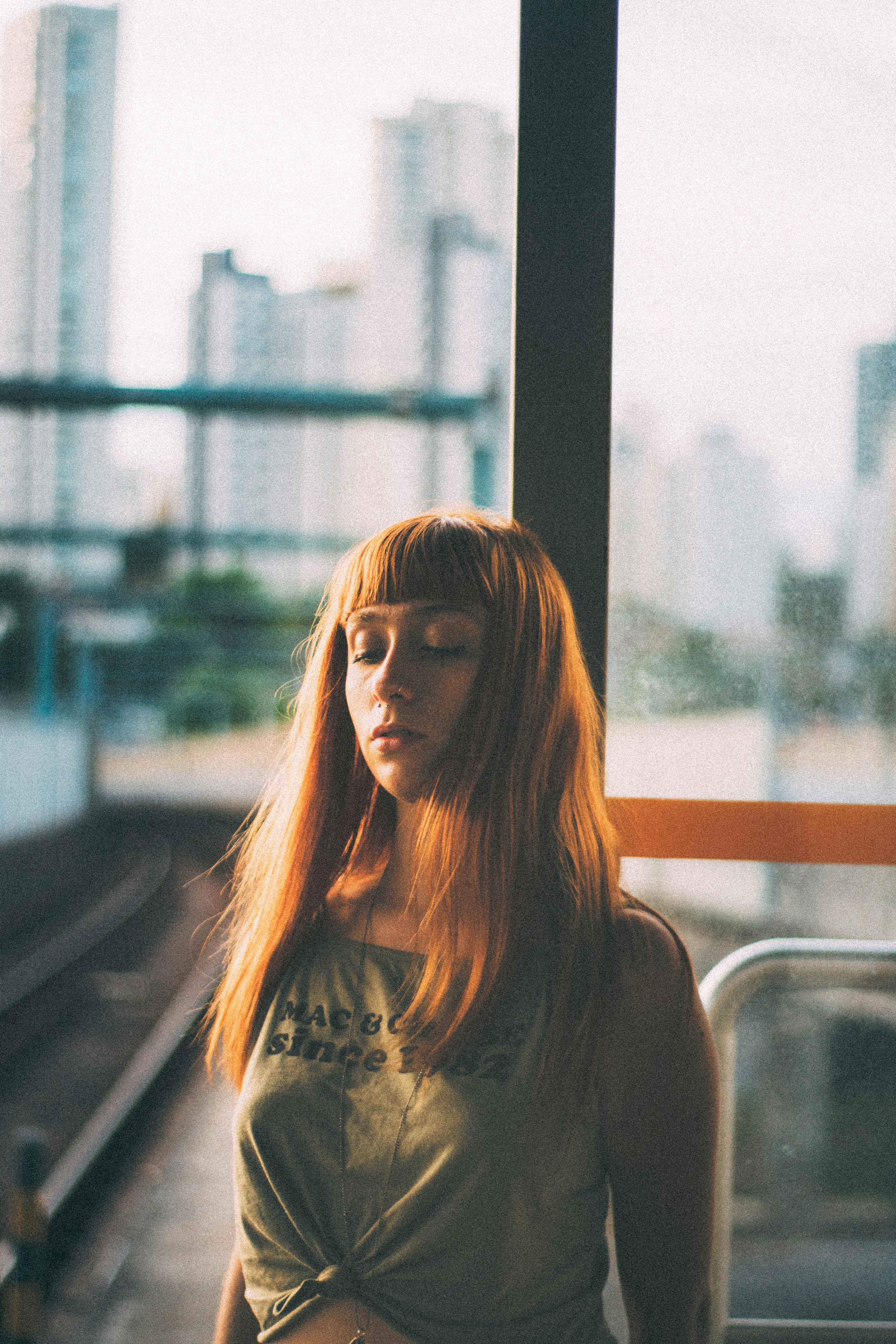 A young woman stands peacefully by a window with an urban backdrop, creating a serene and stylish scene.