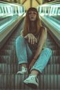From below full body calm young female in jeans sitting on going up escalator and looking at camera