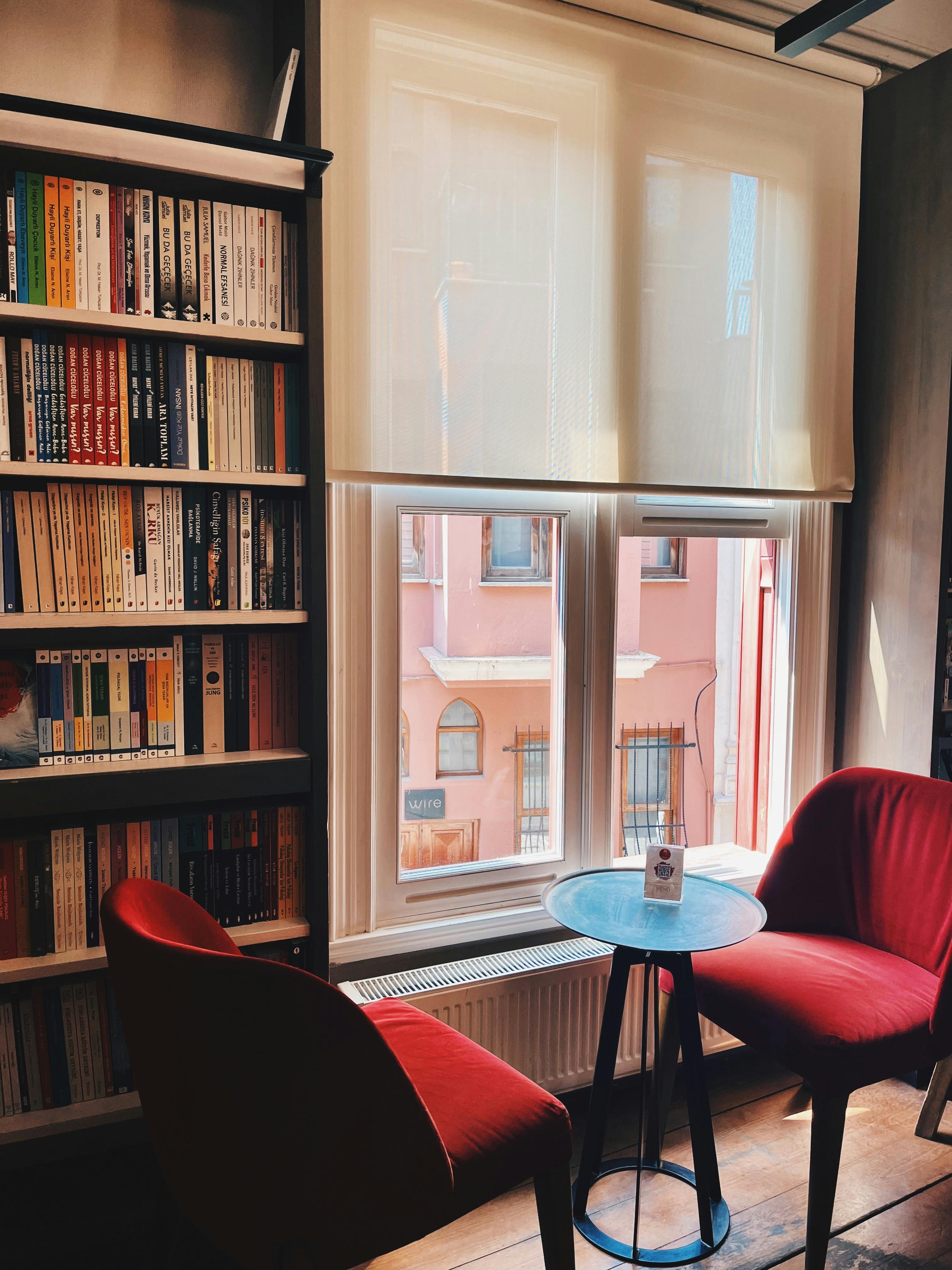 Gratuit Un colț de lectură confortabil cu scaune roșii lângă o fereastră luminată de soare într-o bibliotecă. Fotografie de stoc