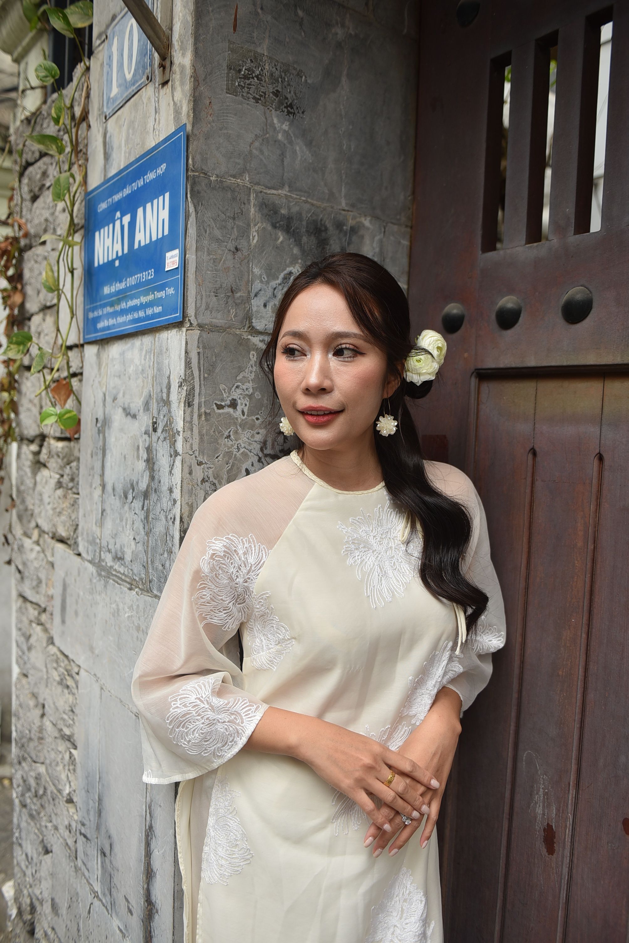 Elegant woman in traditional attire leaning against a stone wall with a sign in Vietnamese.