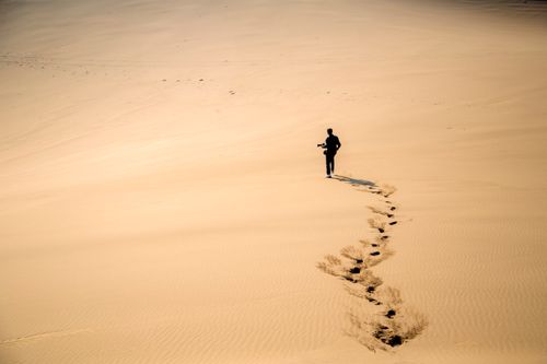 Free A photographer walking across vast sand dunes in the Iranian desert, leaving footprints behind. Stock Photo