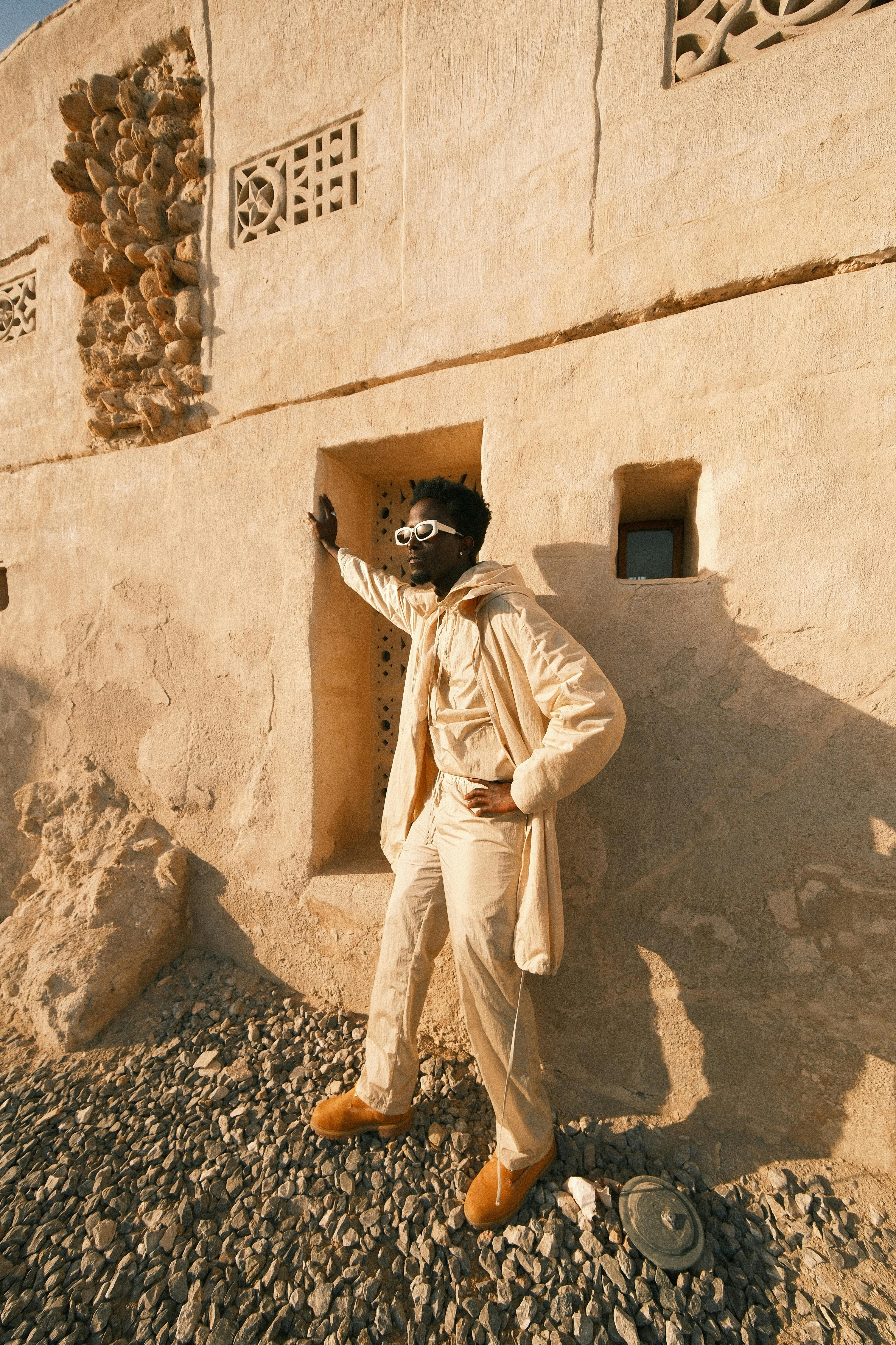 A fashionable man in a beige outfit poses against an ancient, rustic wall.