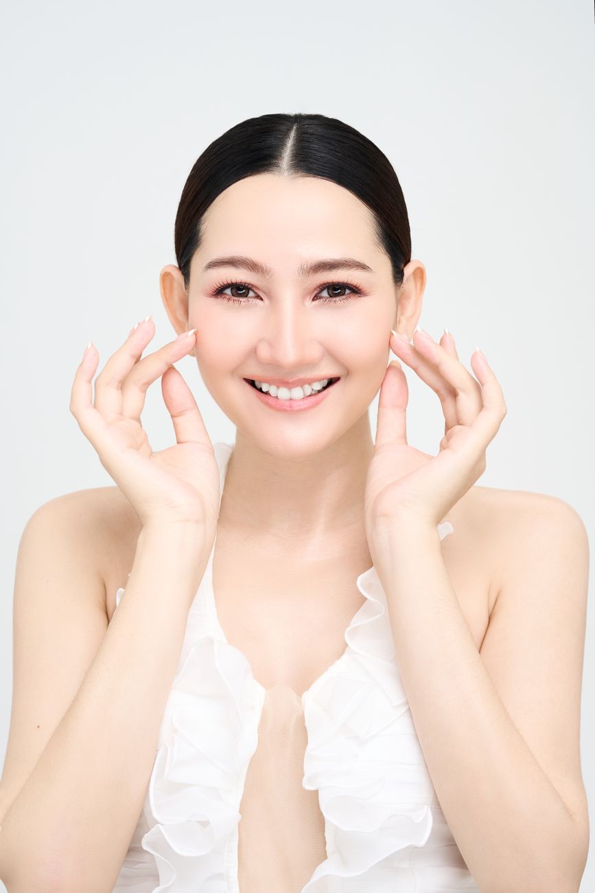 Smiling Asian woman in a white dress applies skincare, showcasing radiant complexion and clear skin.