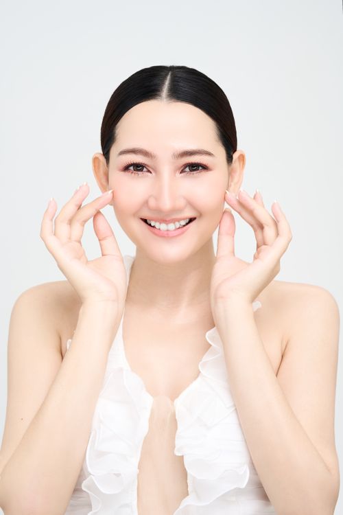 Free Smiling Asian woman in a white dress applies skincare, showcasing radiant complexion and clear skin. Stock Photo