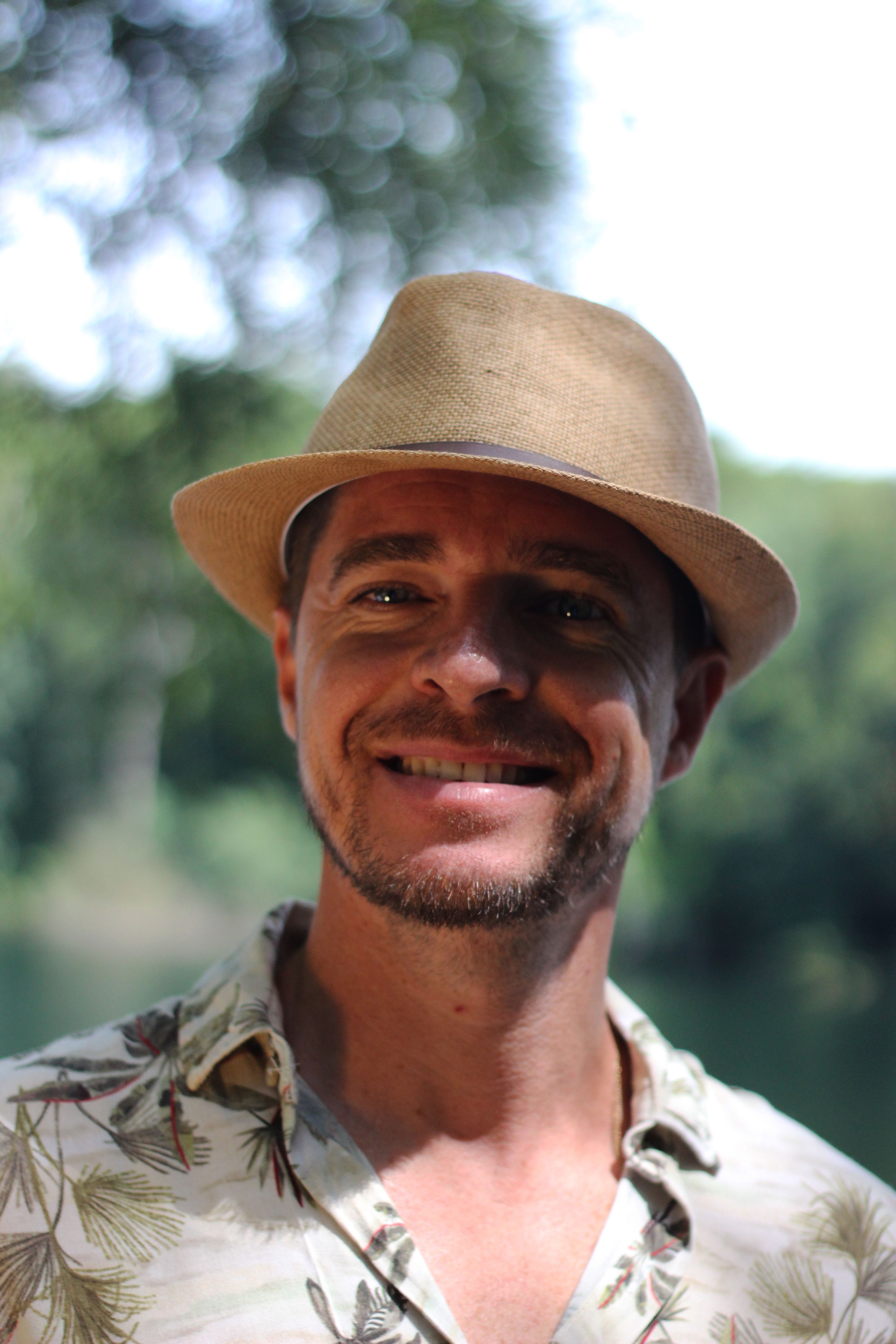 A cheerful man wearing a fedora smiles outdoors amidst greenery in Braga, Portugal.