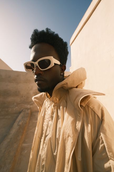 A fashionable man in sunglasses and beige outfit posing against a minimalistic urban backdrop.