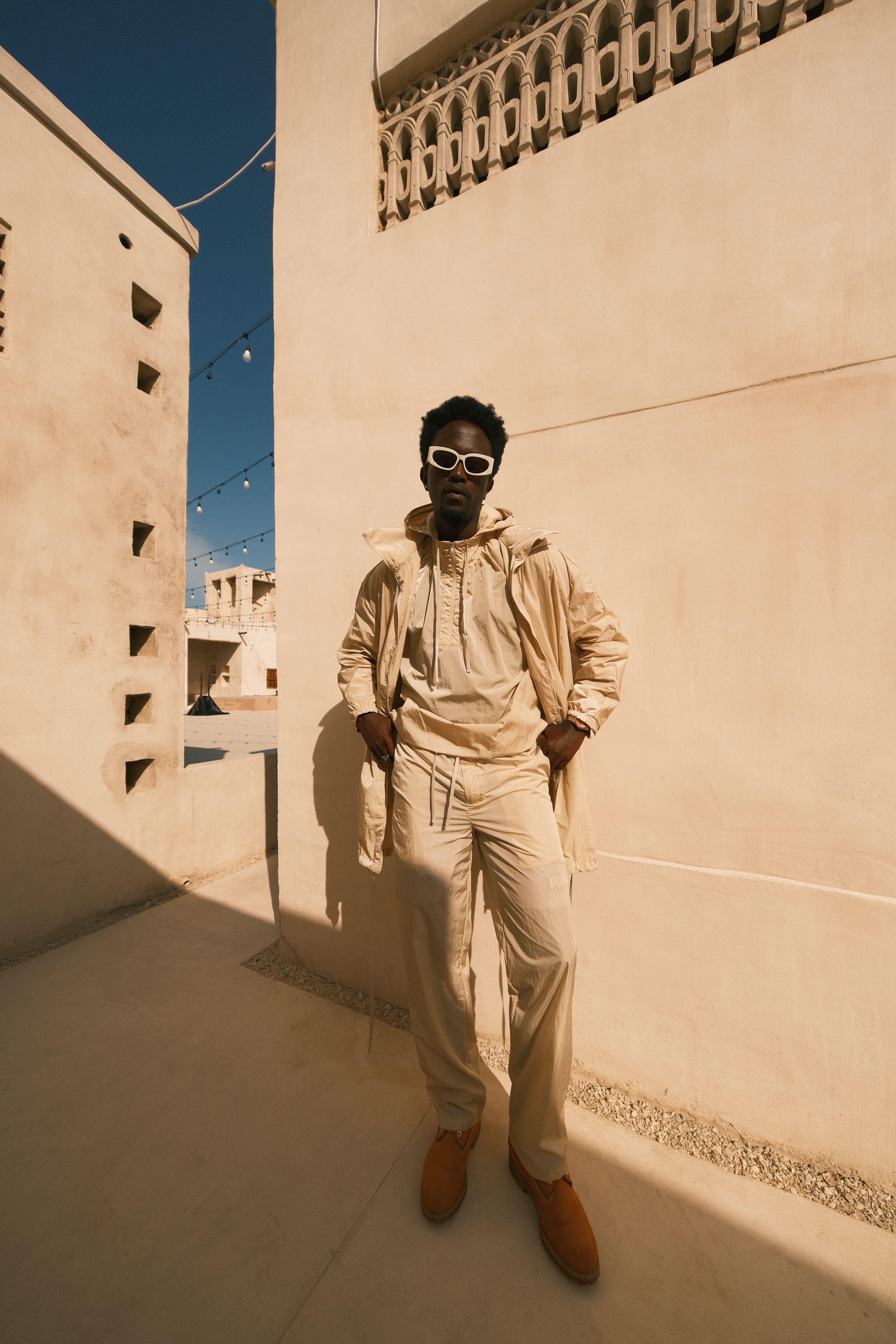 Stylish man in beige outfit and sunglasses posing in a sunlit urban area.