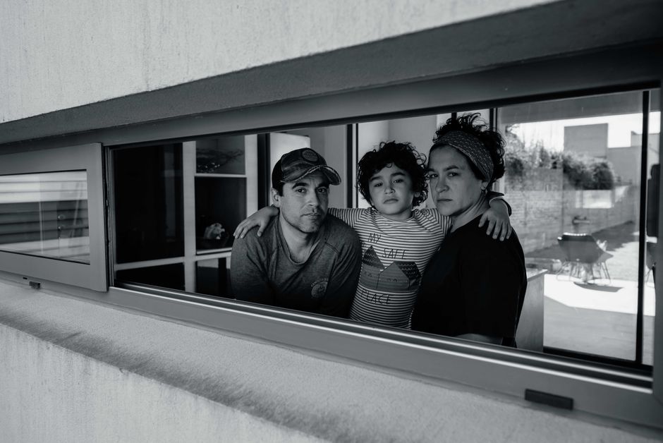 Un retrato en blanco y negro de un trío familiar mirando desde el marco de una ventana, capturando un momento reflexivo.