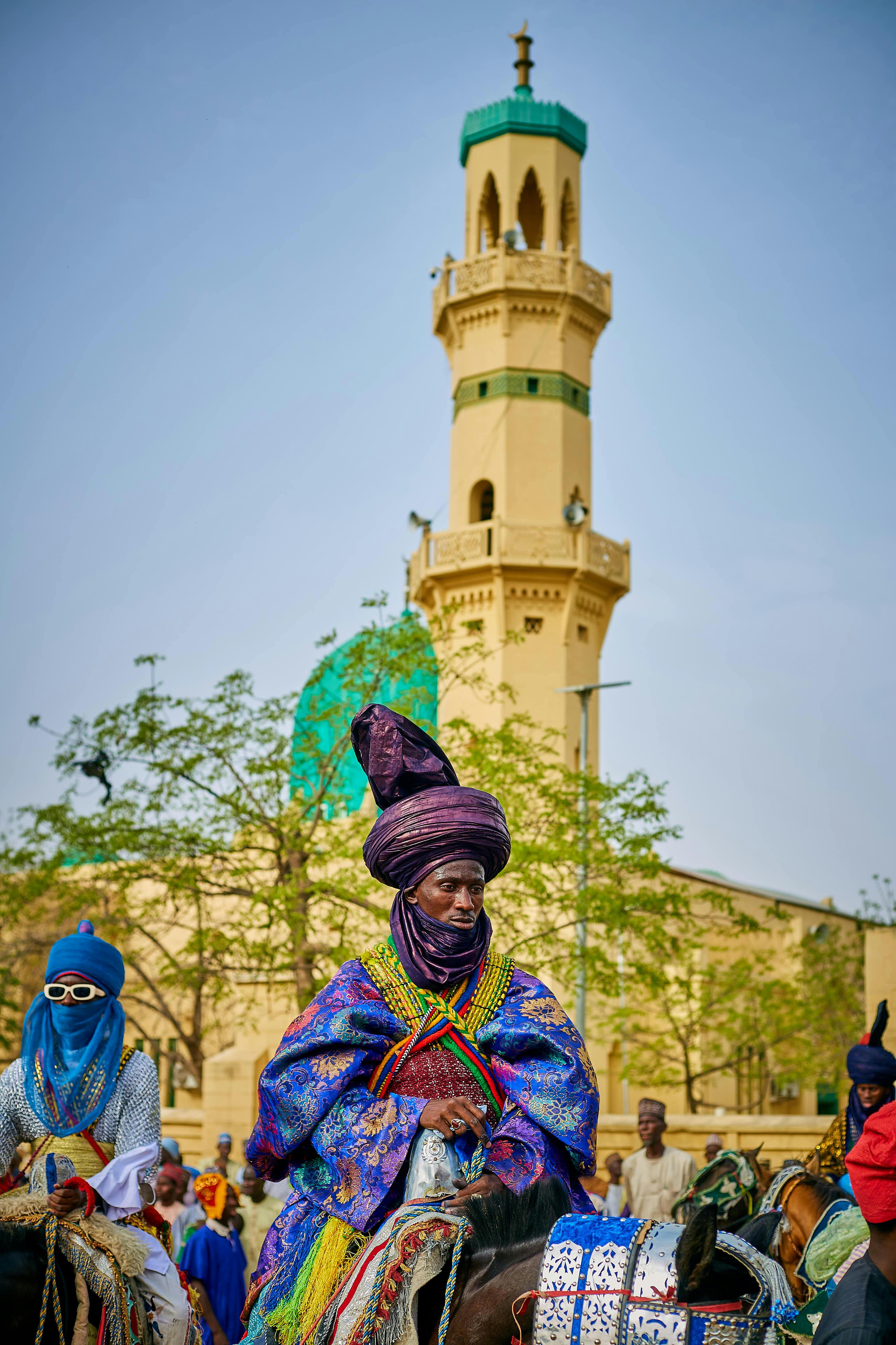 Vibrant traditional horse riders in colorful attire near a mosque tower in Nigeria.