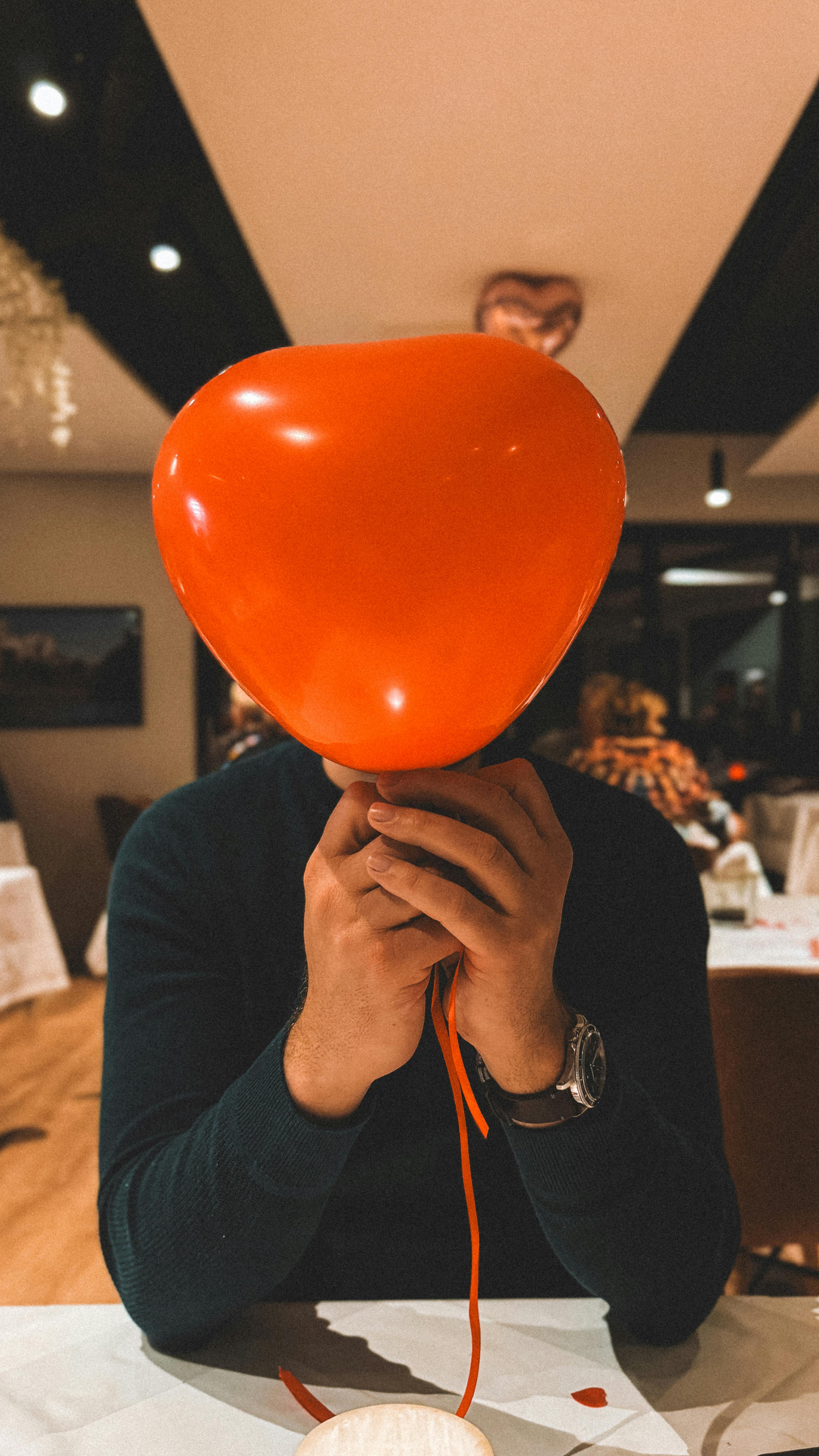 A person playfully holding a heart-shaped balloon at a dining table.