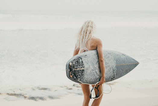 An adult woman carrying a surfboard walks towards the ocean on a sunny day in Bali, Indonesia.