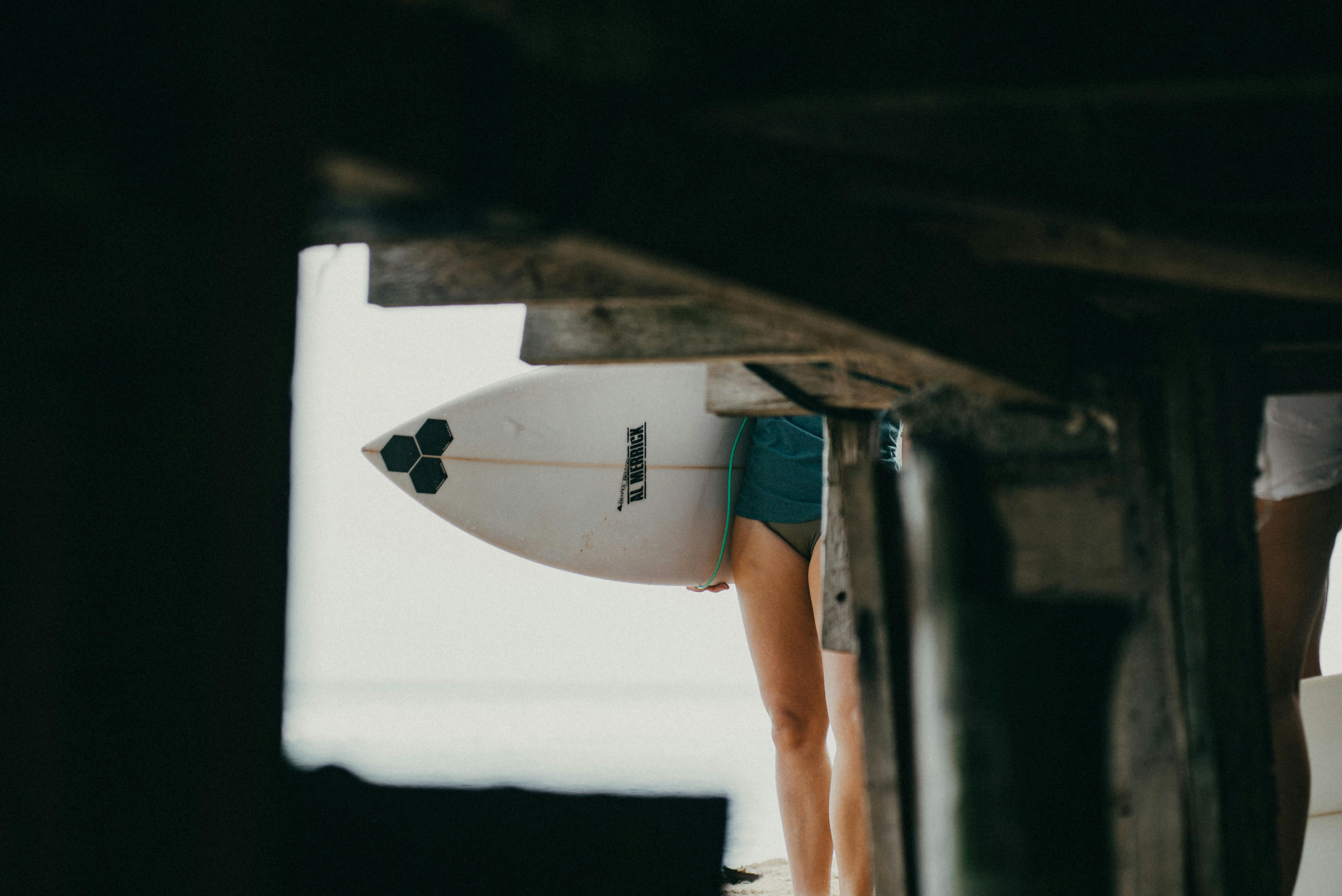 grátis Surfista com prancha sob um píer em Bali, criando uma perspectiva única da vida na praia. Foto profissional
