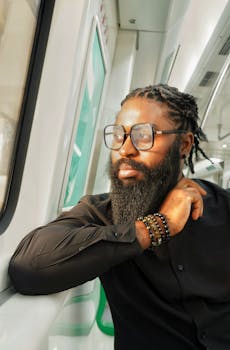 A thoughtful man in a black outfit with a beard and glasses sitting on a train in Abuja, Nigeria.