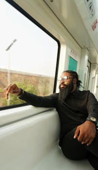 A contemplative man with dreadlocks and glasses looking out of a train in Abuja, Nigeria, during the day.
