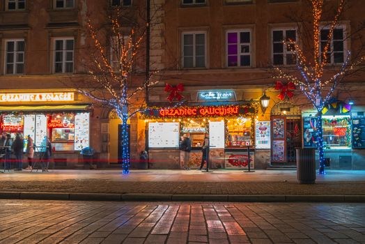 Festive night lights illuminate a Warsaw street with Christmas decorations and shops.